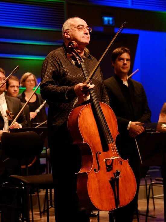 Alexander Kaganovsky on stage with his cello and bow, standing in front of the orchestra